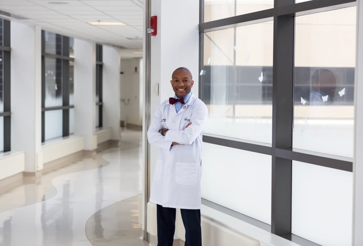 Dr. Bignall is wearing a white coat and bow tie. His arms are crossed. He's smiling and standing in a hallway with bright windows behind him.