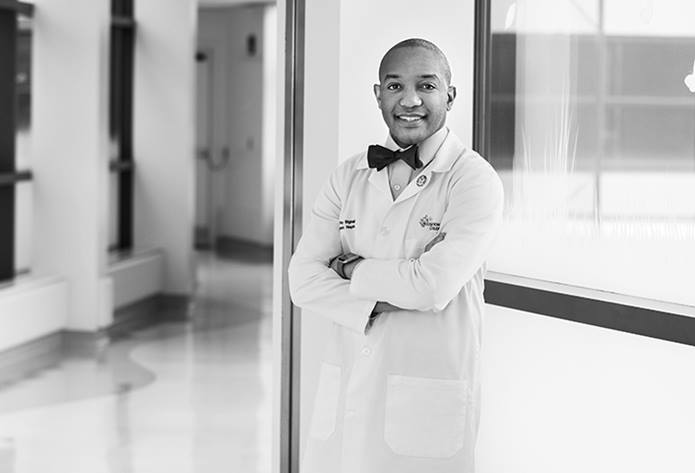 Dr. Bignall is wearing a white coat and bow tie. His arms are crossed. He's smiling and standing in a hallway with bright windows behind him.