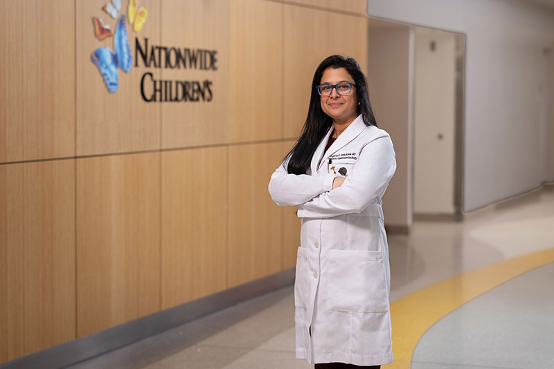 Physician in a white lab coat standing with arms crossed in a Nationwide Children’s hospital hallway, with the Nationwide Children’s logo displayed on the wall behind her.