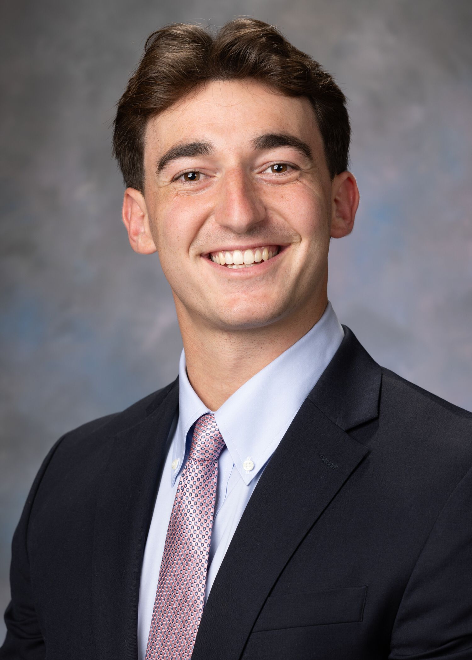 Portrait of Timothy Dillon. Timothy is wearing a blue dress shirt, purple tie, and a dark colored suit jacket.