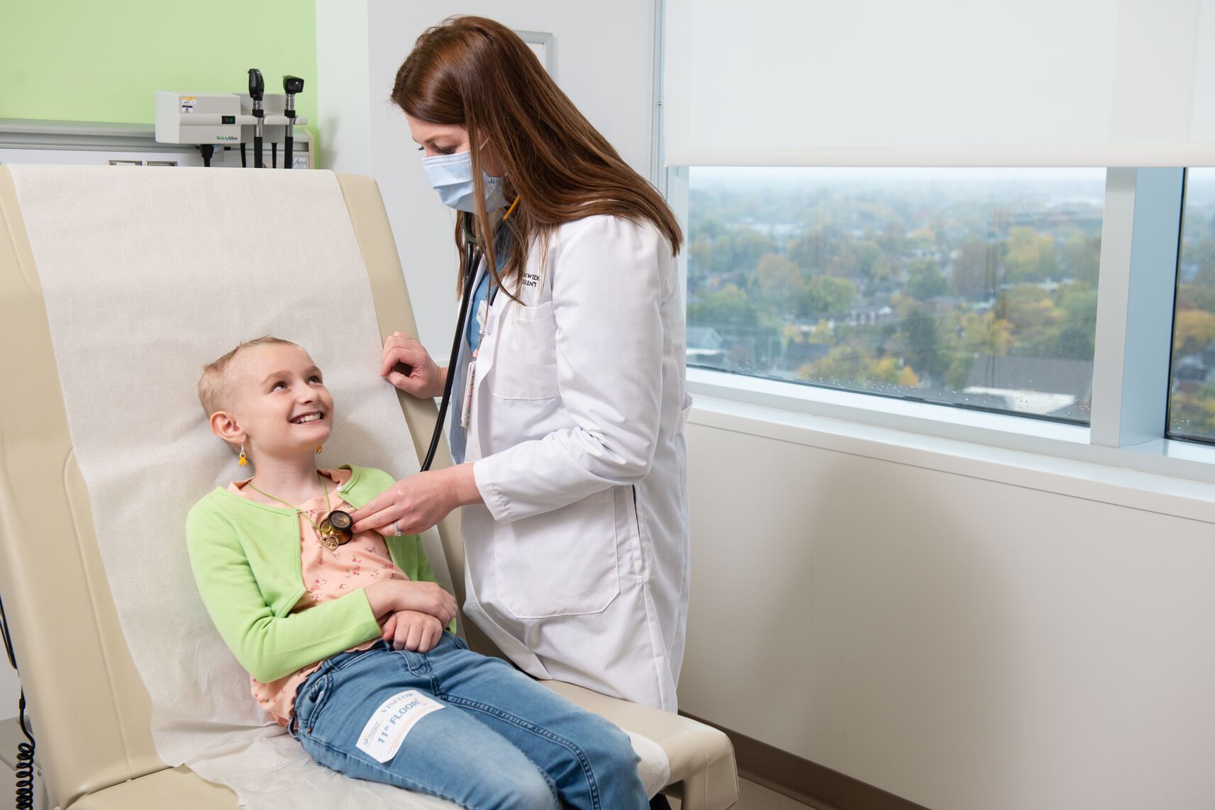 A healthcare professional wearing a face mask and white coat uses a stethoscope to listen to the chest of a child seated on an exam table in a medical room, with diagnostic equipment on the wall and a window showing an outdoor view in the background.