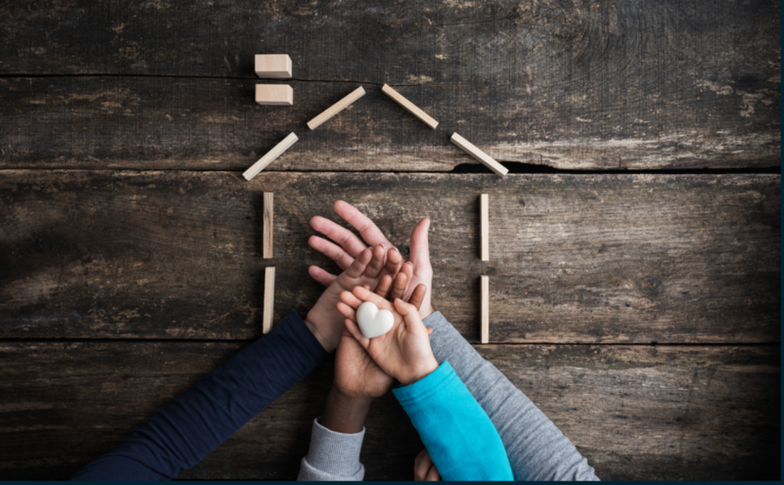 Four hands stacked on top of each other with the smallest hand at the top, holding a rock shaped like a heart. There is an outline of a house made out of blocks in the background, lying on a wooden plank background.