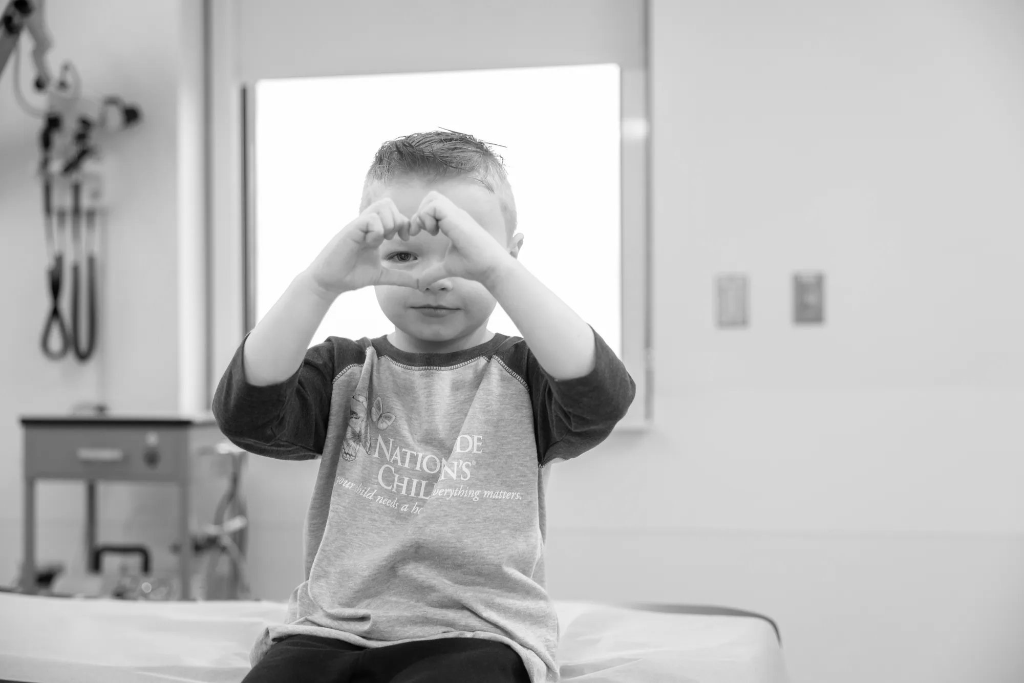 A young boy is sitting on the exam table in the hospital. He is using his hands to make a heart symbol in front of his face.