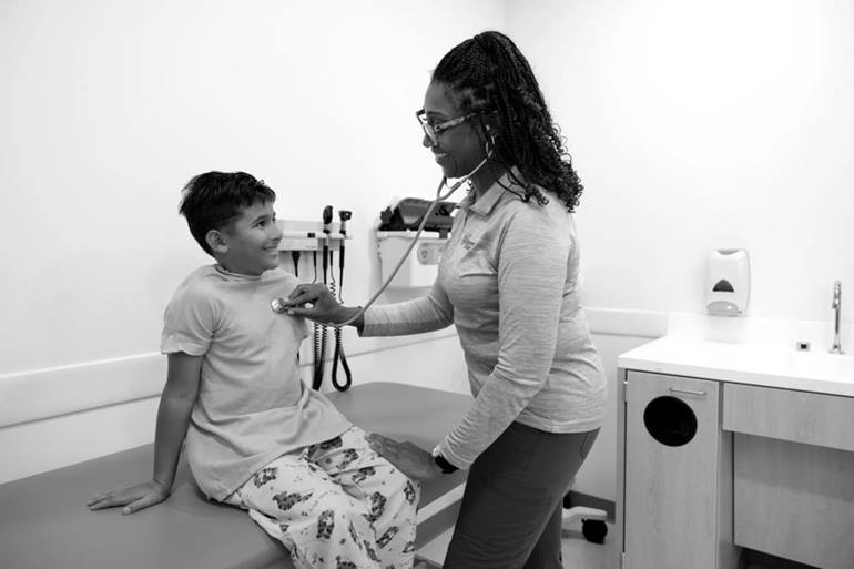 A provider is holding a stethoscope to a patient's chest while he sits on the examination table. They are both smiling.