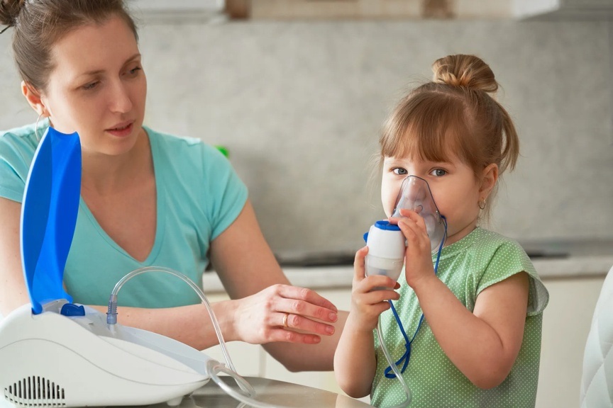A young girl wearing a green shirt is holding an inhaler to her face while a provider is helping her.