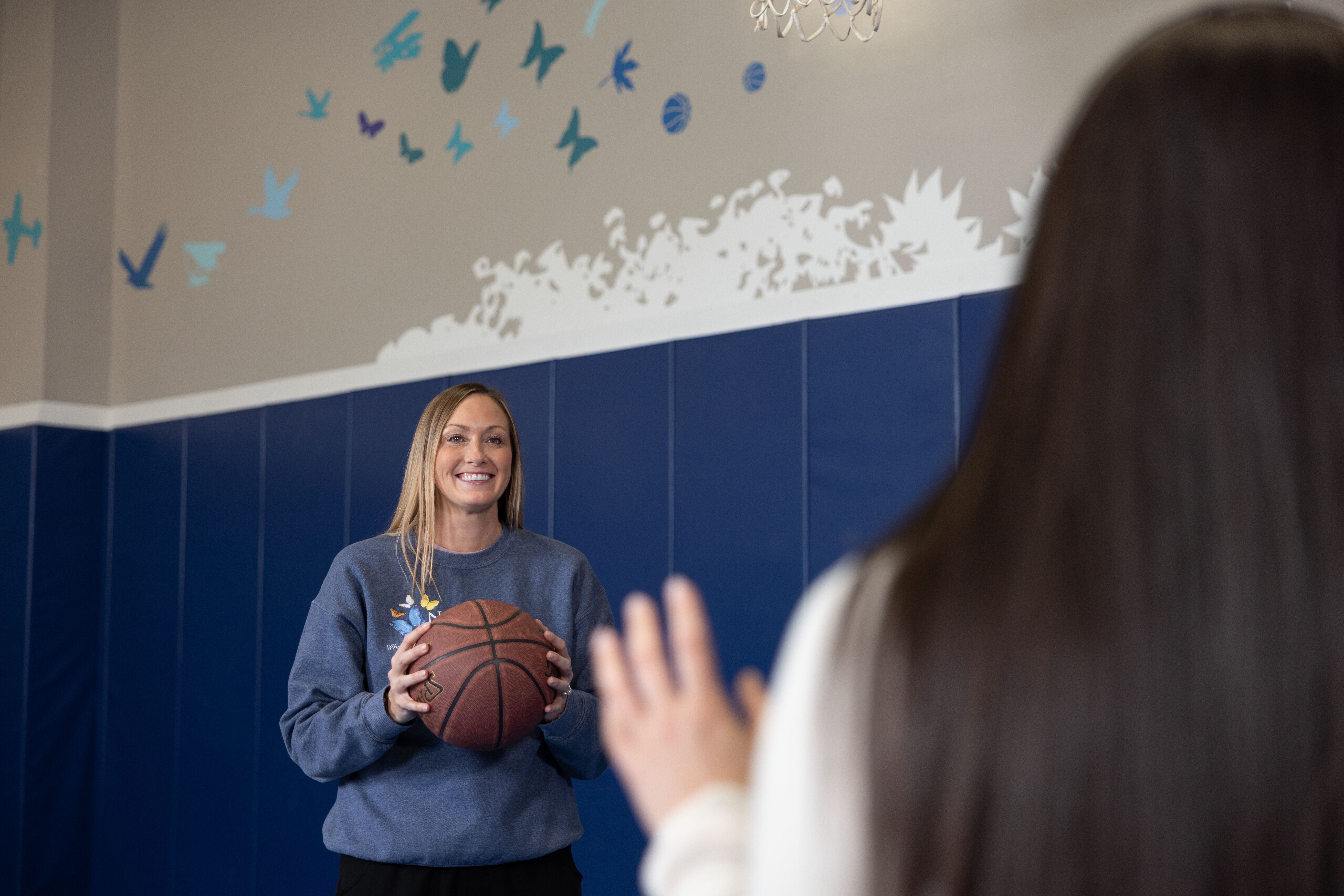 A young lady in a grey sweatshirt is holding a basketball and looking to pass to another person who has dark hair near the camera. 