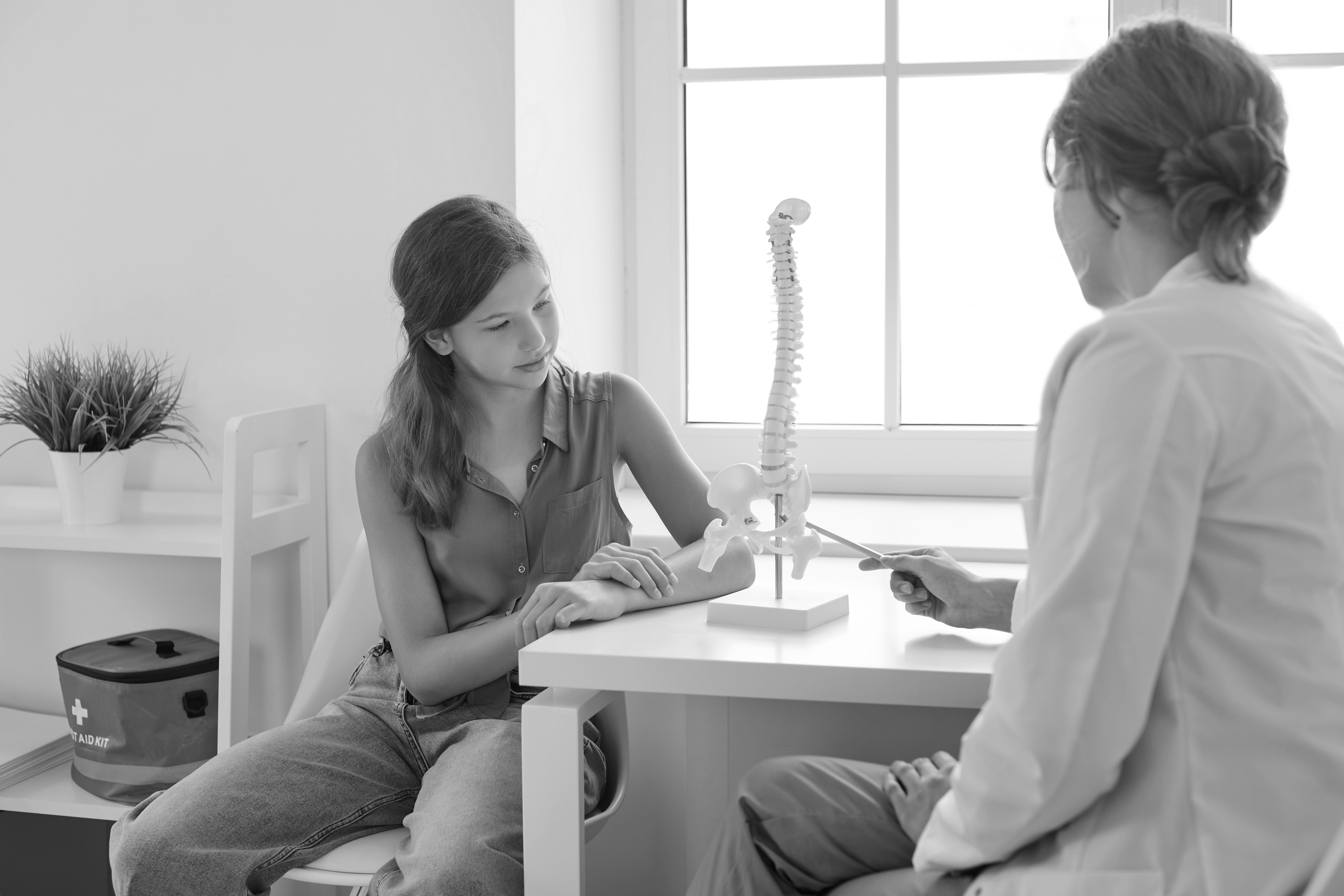 A young girl sits at a doctor’s office desk, listening as a healthcare professional explains something using a model of the human spine. The girl looks attentive, resting her arm on the table, while the doctor gestures toward the spine model with a pointer.