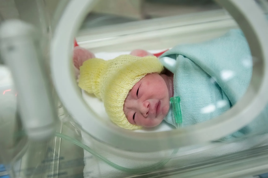 Infant swaddled in a hospital bassinet with a yellow hat on.