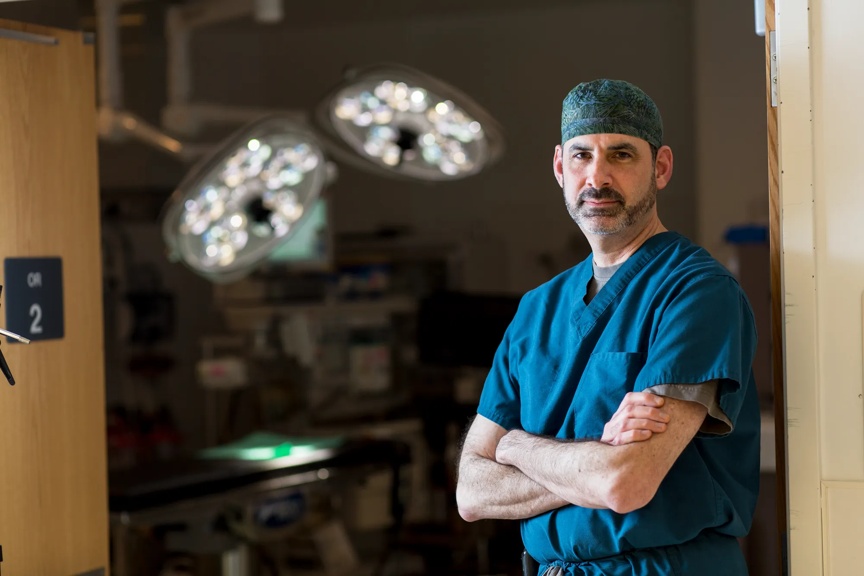 Male surgeon in scrubs standing in front of an operating room.