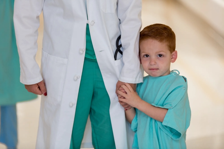 Young boy standing in scrubs holding a doctor's hand in a hospital hallway.