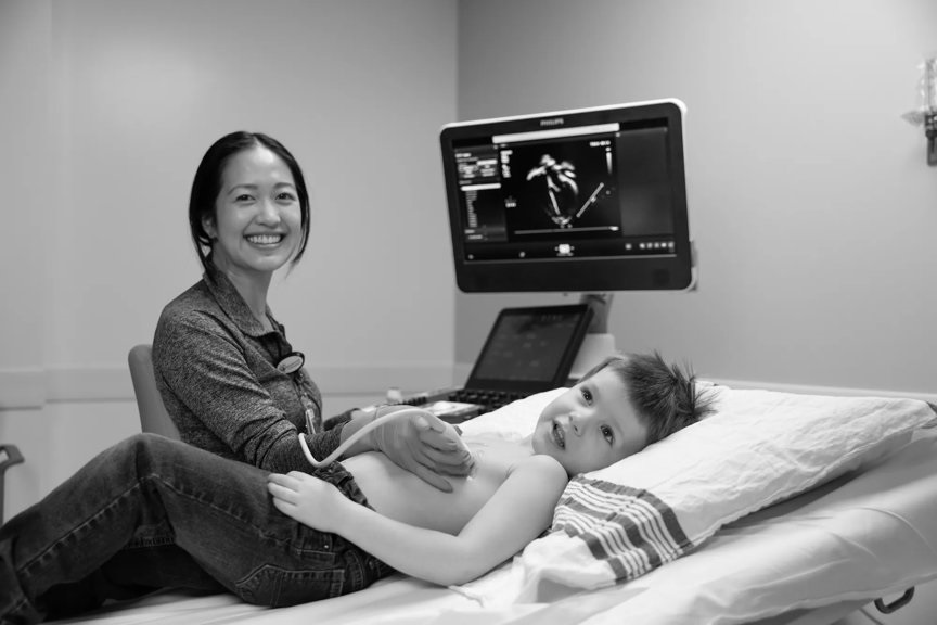 Provider is smiling at the camera which checking the chest and lungs of a young patient on the exam table. There is a monitor behind both of them.