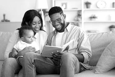 Two parents sitting on a couch reading a book to their baby, smiling.