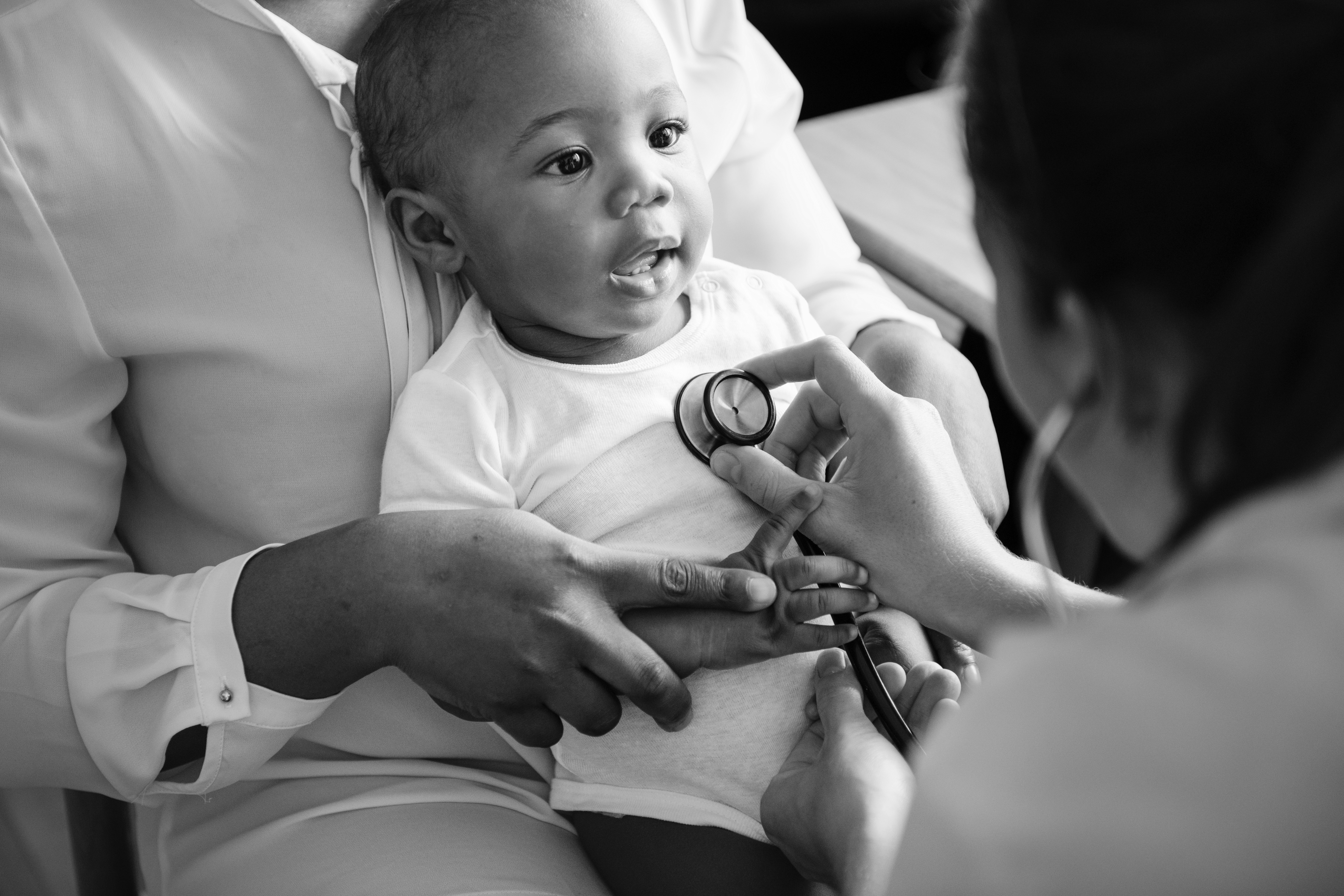 Healthcare professional using a stethoscope to listen to the chest of a baby who is sitting on an adult's lap during a medical examination.