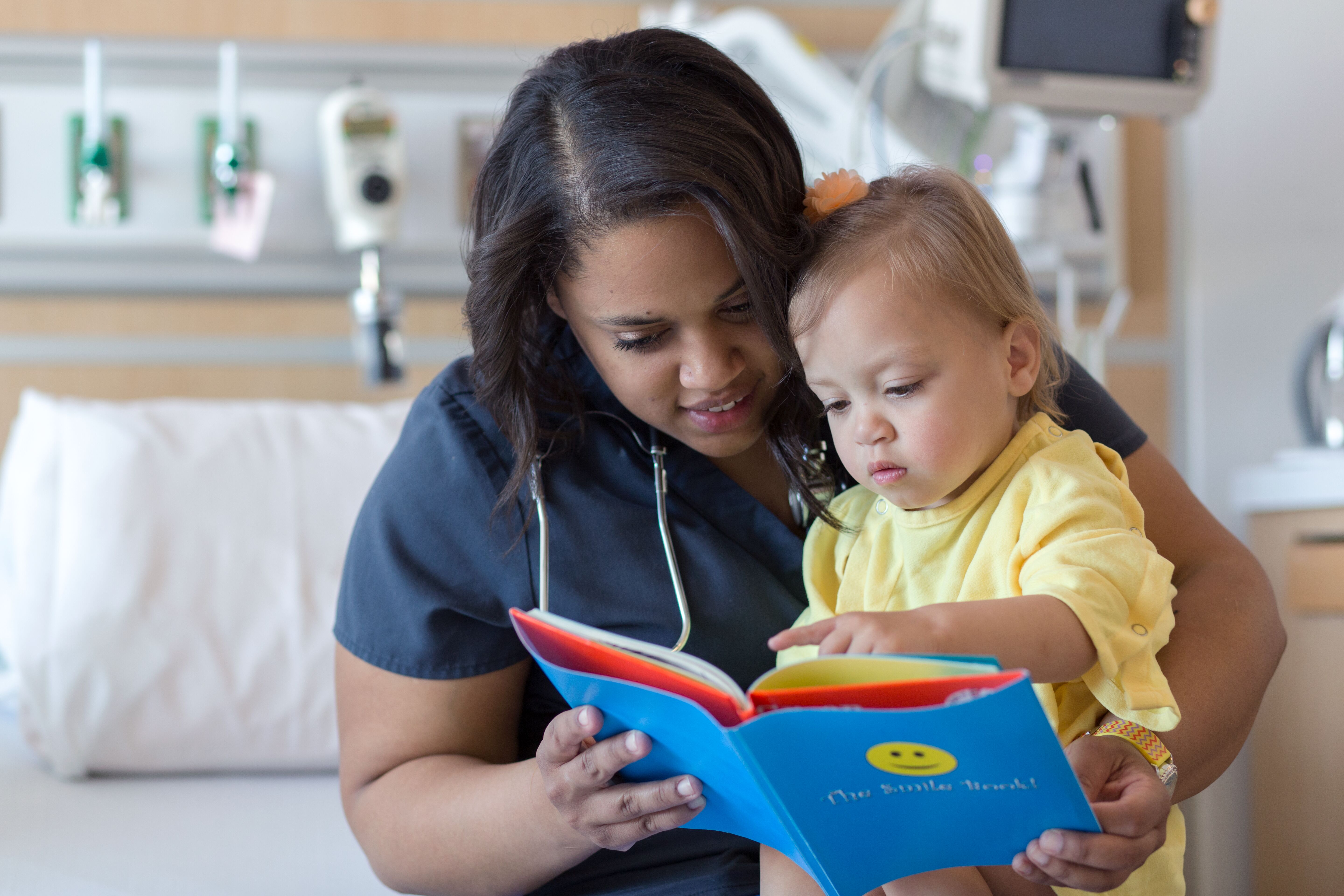 A provider who is wearing a blue shirt is reading a blue book with a little girl wearing a yellow shirt. There is a hospital bed in the background.