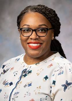 Portrait of Mattina Davenport, woman with long braided brown hair, glasses, and a patterned white blouse.