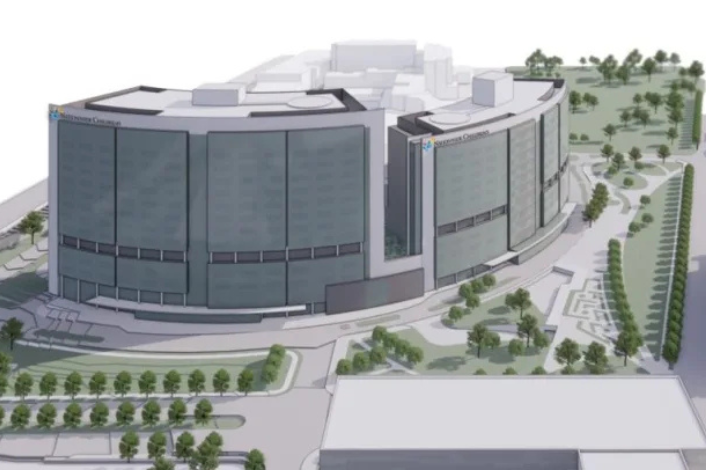 Architectural rendering of a large, modern hospital tower and surrounding campus viewed from an elevated angle. The multi-story curved building features expansive glass façades, light gray exterior panels, and multiple connected wings. Landscaped grounds surround the facility, including rows of trees, walking paths, green spaces, plazas, and drop-off areas. Additional buildings are visible adjacent to the tower, with roads and sidewalks connecting the campus. The rendering appears to depict a planned healthcare expansion or new patient tower integrated into an urban medical center setting.