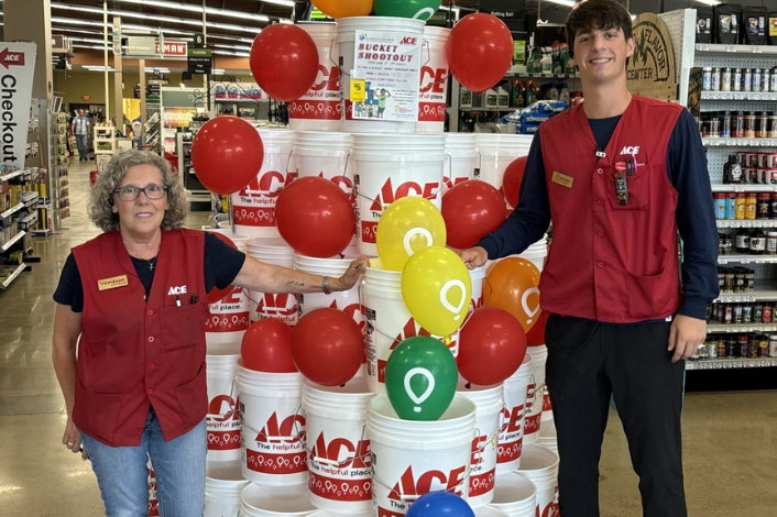Two ACE Hardware employees wearing red store vests stand on either side of a large display made from stacked white ACE-branded buckets decorated with red, yellow, green, orange, and blue balloons. A sign at the top of the display reads “Bucket Shootout,” and store aisles and merchandise are visible in the background.
