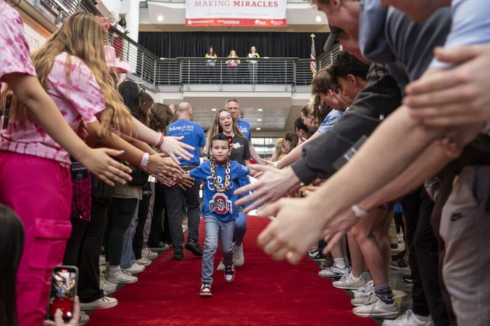 A child wearing a blue Ohio State-themed shirt and oversized gold chain walks down a red carpet through a crowd of people lined up on both sides with outstretched hands. The event takes place inside a large atrium, where a banner reading “Making Miracles” hangs above a balcony in the background.