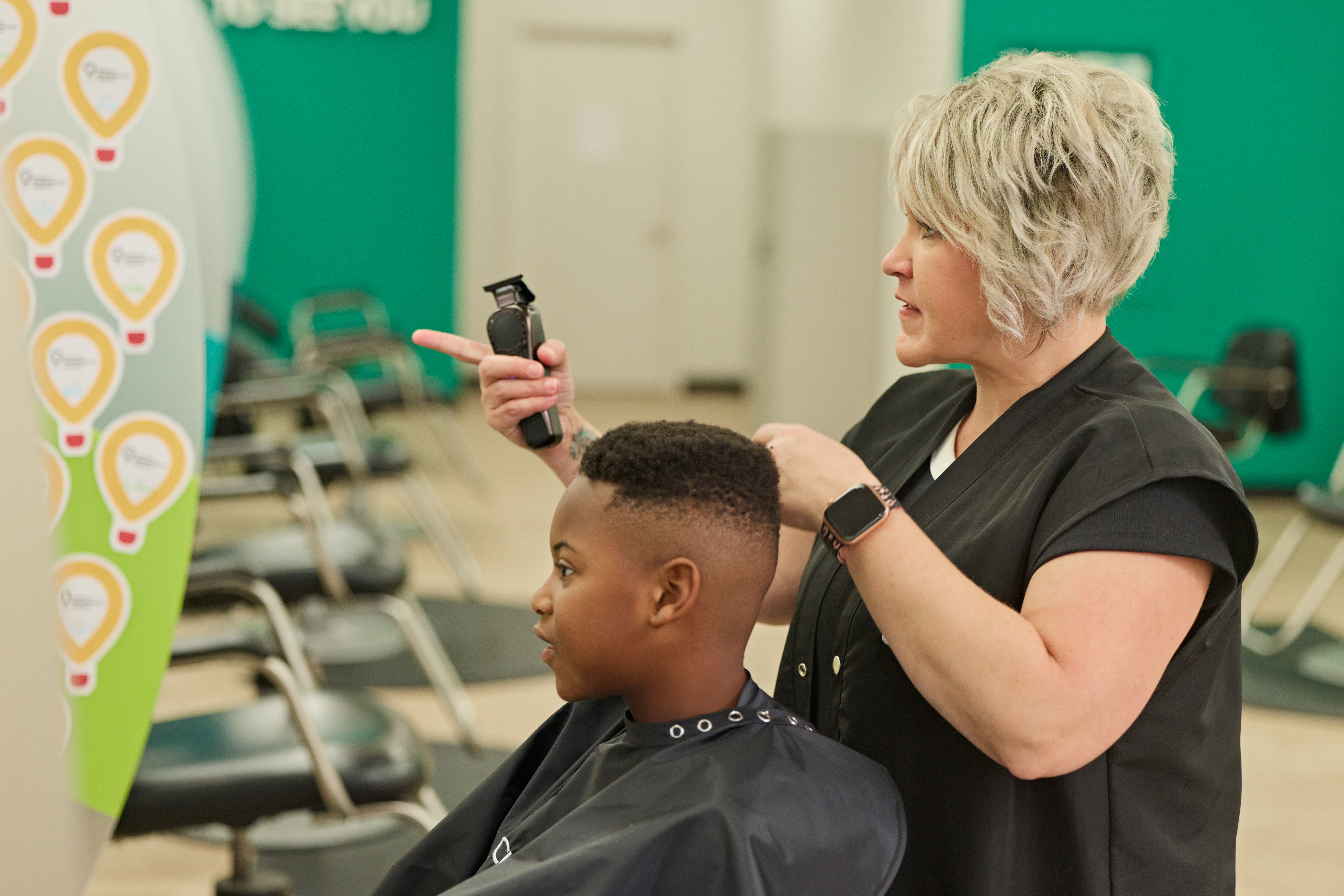 A hairstylist uses electric clippers to cut a child’s hair inside a salon. The child sits in a barber chair wearing a protective cape while the stylist works on a short fade haircut. Salon chairs and mirrors are visible in the background, along with a Children’s Miracle Network Hospitals-themed display featuring balloon-shaped icons.