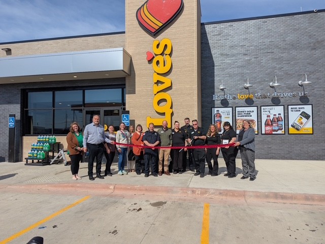 A group of people stands outside a Love’s Travel Stop store during a ribbon-cutting ceremony. The group holds a long red ribbon across the entrance, with the large yellow Love’s sign mounted on the building behind them. Store promotional signs are displayed on the exterior wall, and the parking lot is visible in the foreground.
