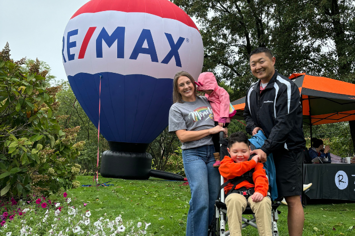 Two adults and two children pose outdoors on a grassy area in front of a large RE/MAX hot-air-balloon-shaped display. One child sits in a wheelchair wearing an orange jacket, while another child in a pink jacket is being held. Trees, flowers, and an event tent are visible in the background.