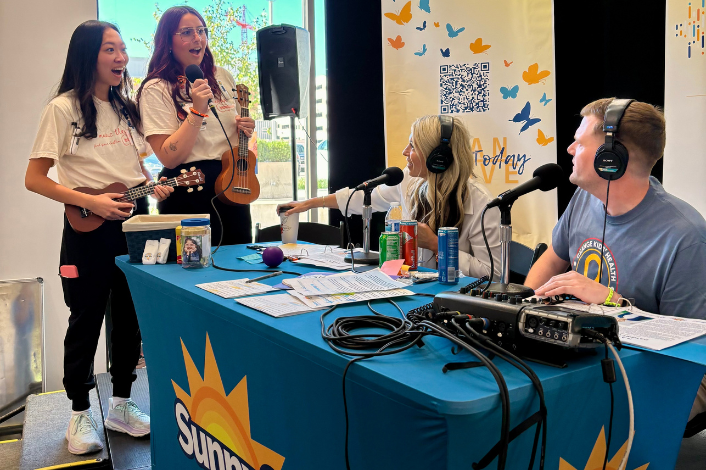 Two young musicians stand beside a live radio broadcast table, each holding a ukulele while one speaks into a microphone. Two radio hosts wearing headsets sit behind the table with microphones, audio equipment, and notes in front of them. A banner with butterflies and a QR code stands in the background, and the event takes place indoors near large windows.