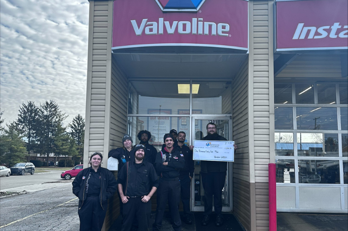 A group of Valvoline Instant Oil Change employees stands outside a service center beneath a large Valvoline sign. One employee holds an oversized ceremonial check presented to Nationwide Children’s Hospital, while the rest of the team gathers near the entrance. The service bay doors, parking lot, and trees are visible in the background on an overcast day.