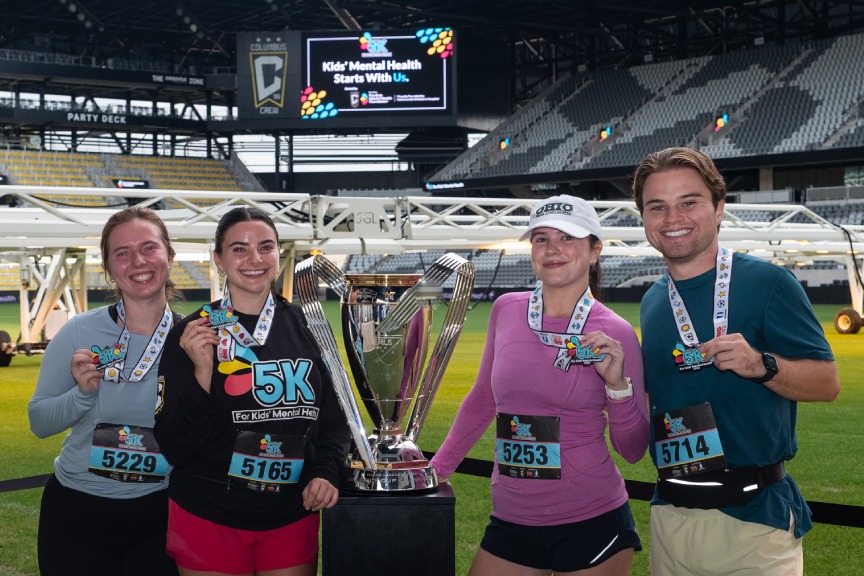A group of four adult runners smile, holding the medals around their necks. A soccer stadium is in the background.