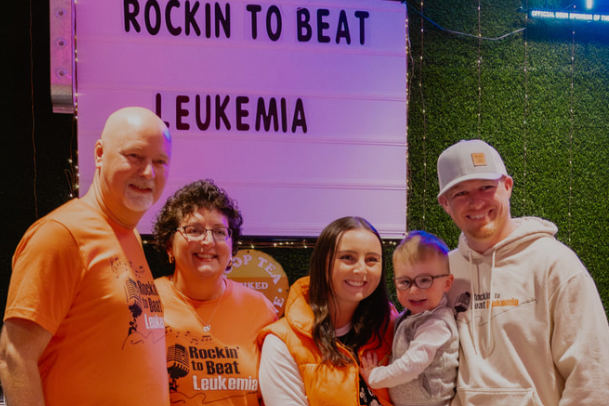 A group of five people standing together indoors in front of a large sign that reads “ROCKIN TO BEAT LEUKEMIA.” Several individuals are wearing bright orange shirts or vests with matching text promoting the event, while one person is holding a small child. The background includes a green textured wall and additional signage, creating a supportive and community-focused atmosphere.