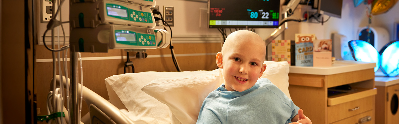 Connor is bald and wearing a blue hospital gown, He's smiling, sitting up in a hospital bed with get well cards behind him.