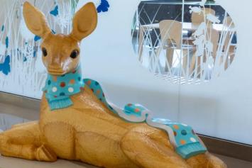 A wooden sculpture of a deer wearing a blue scarf, positioned in a hospital waiting area with decorative wall decals.
