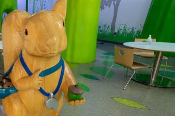 A large wooden squirrel statue stands in a colorful hospital lobby, holding acorns, surrounded by green trees and seating areas.