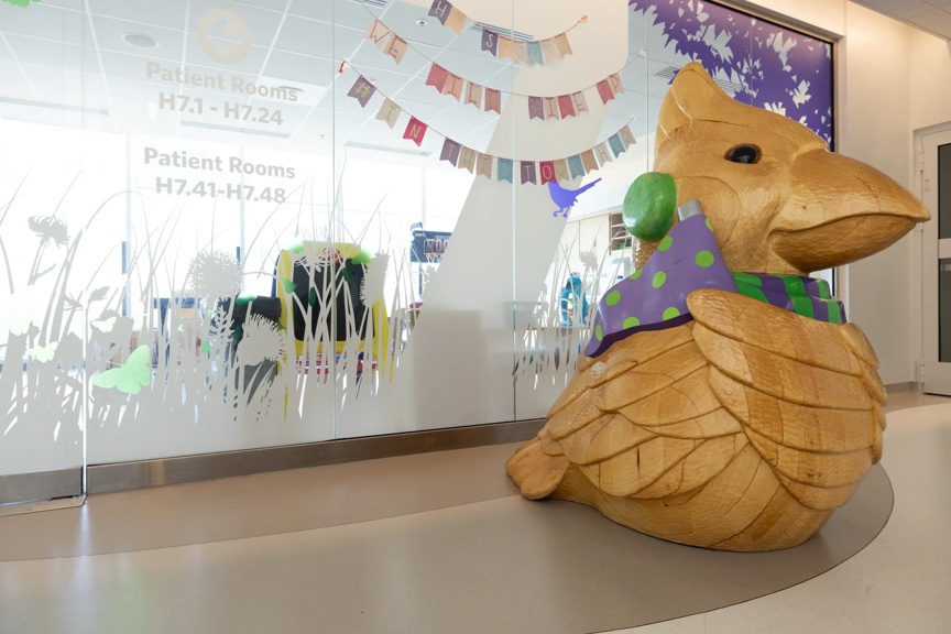 A large wooden bird sculpture in a hospital hallway near patient room signage, adding a welcoming touch to the environment.