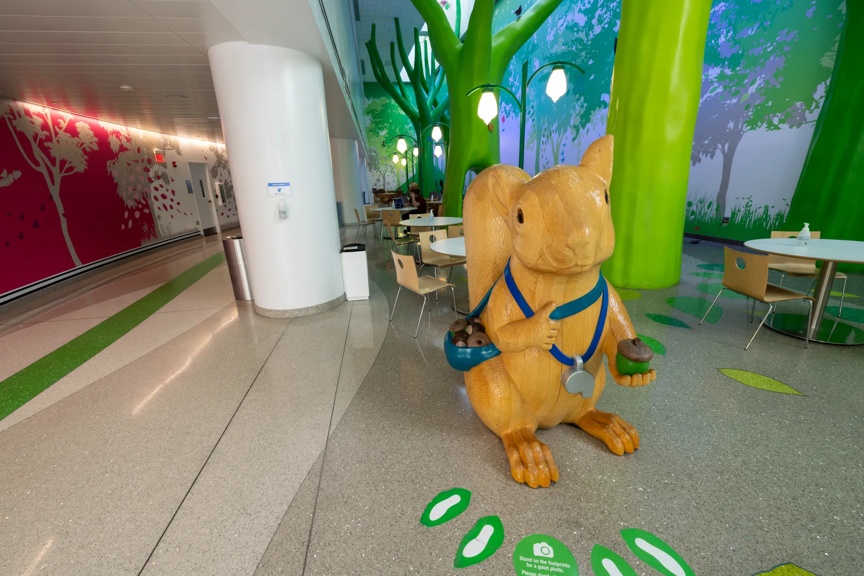 A large wooden squirrel statue stands in a colorful hospital lobby, holding acorns, surrounded by green trees and seating areas.