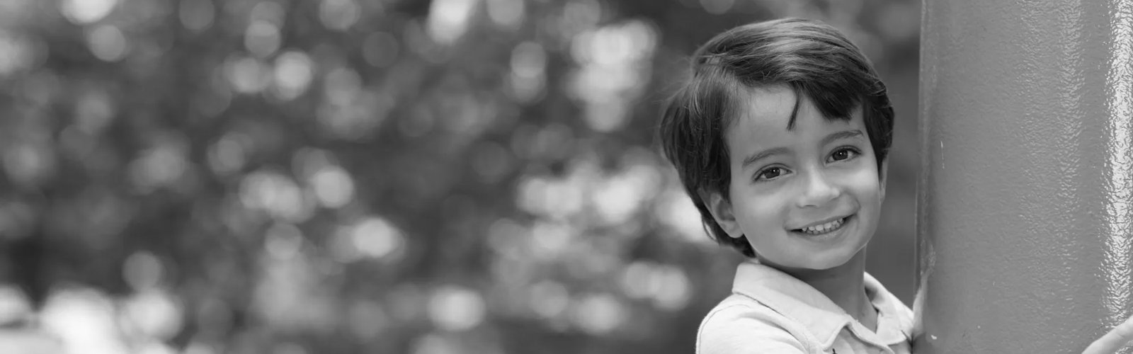 A young, smiling boy leans against a metal column on a summer day. He's wearing a light polo and has dark, short hair.