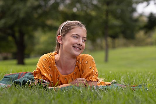 A middle school aged girl rests on a blanket in a park. It's summer and she's smiling, staring off to the distance.