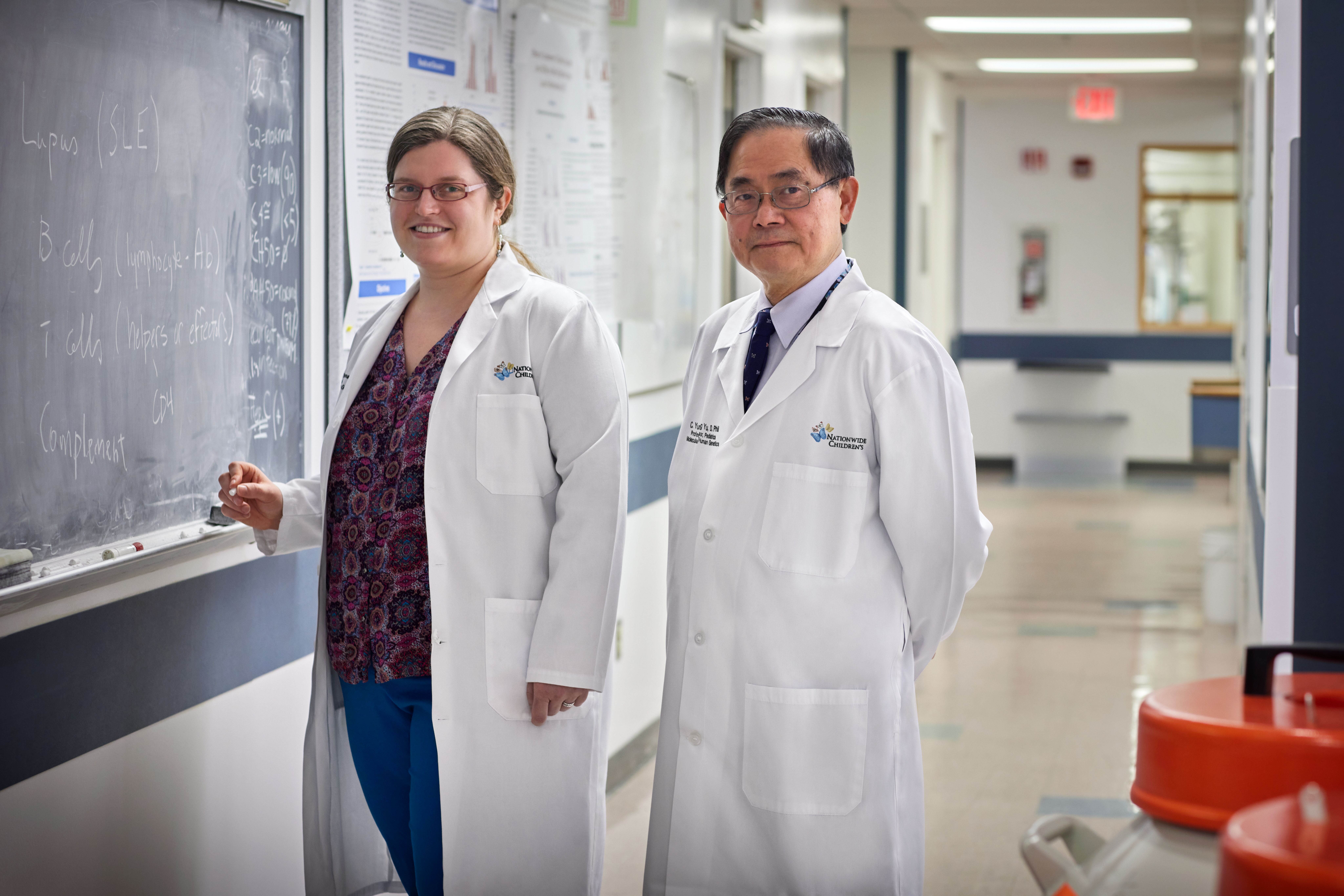 Two medical professionals in white lab coats stand in a hospital corridor, one pointing to a chalkboard with notes.