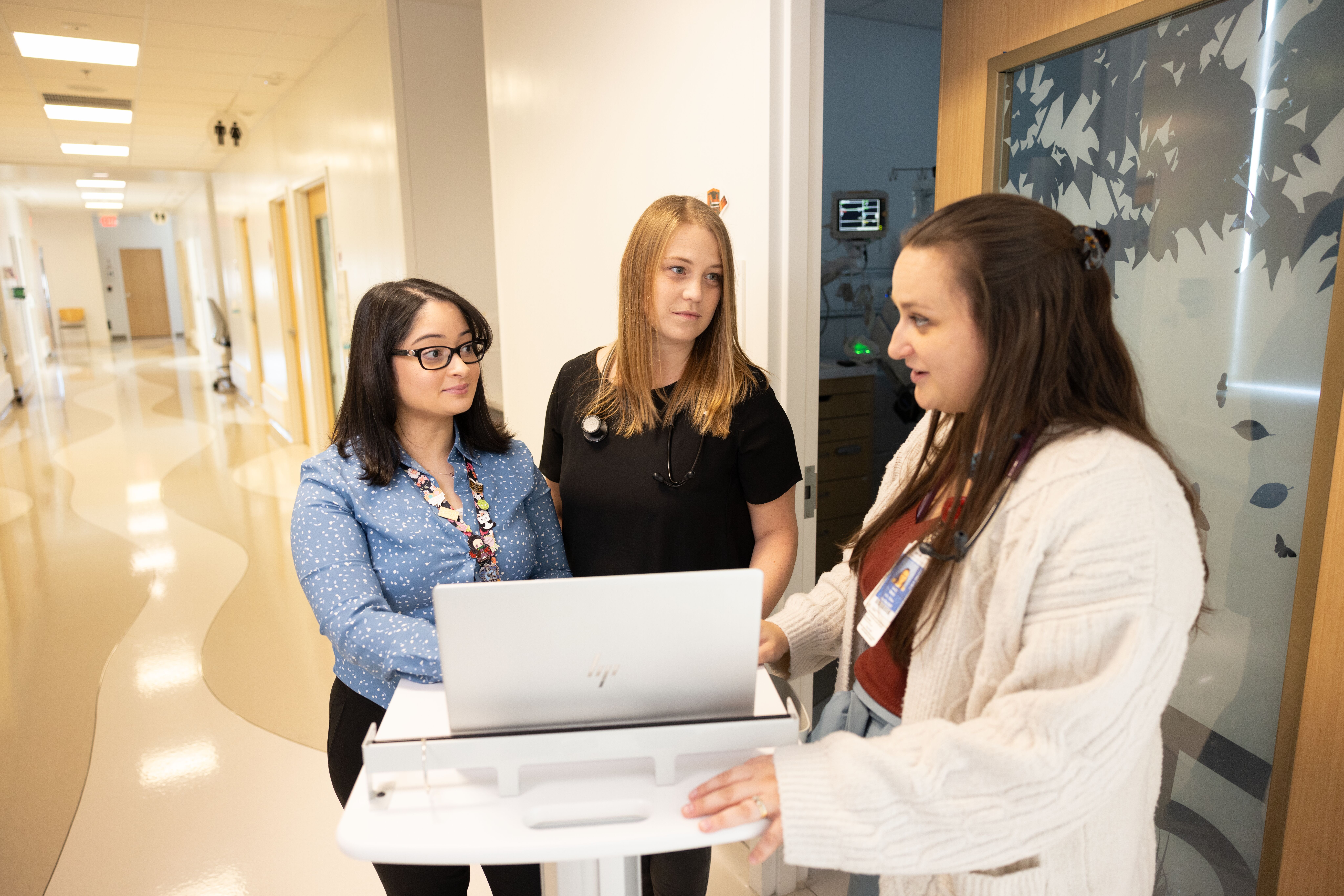 Three healthcare professionals collaborating at a computer station in a hospital corridor.