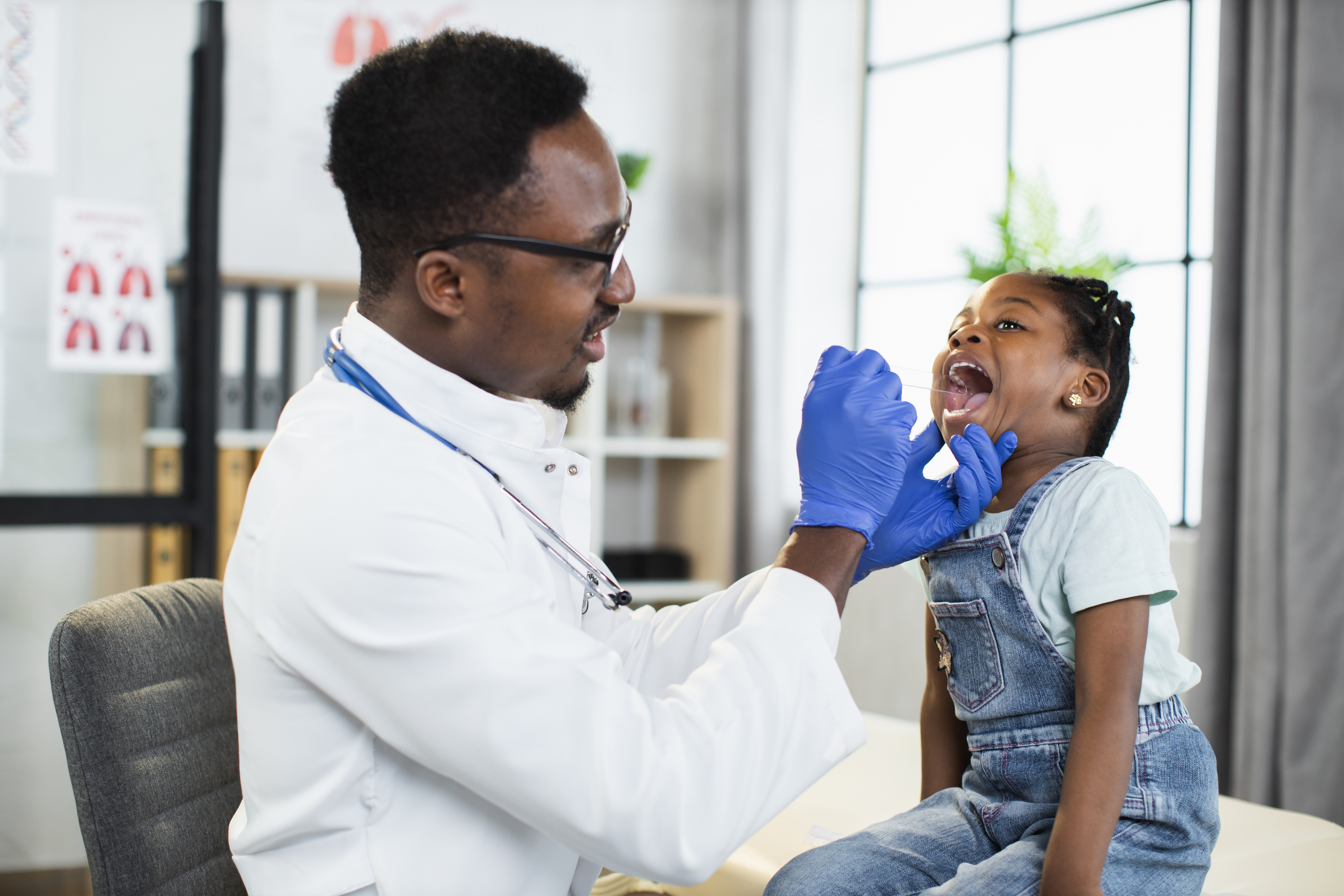 Lovely little afro-american child girl sitting on the couch at doctor's office, while handsome black male doctor examining her throat and mouth with spatula. Pediatrics concept by sofiko14