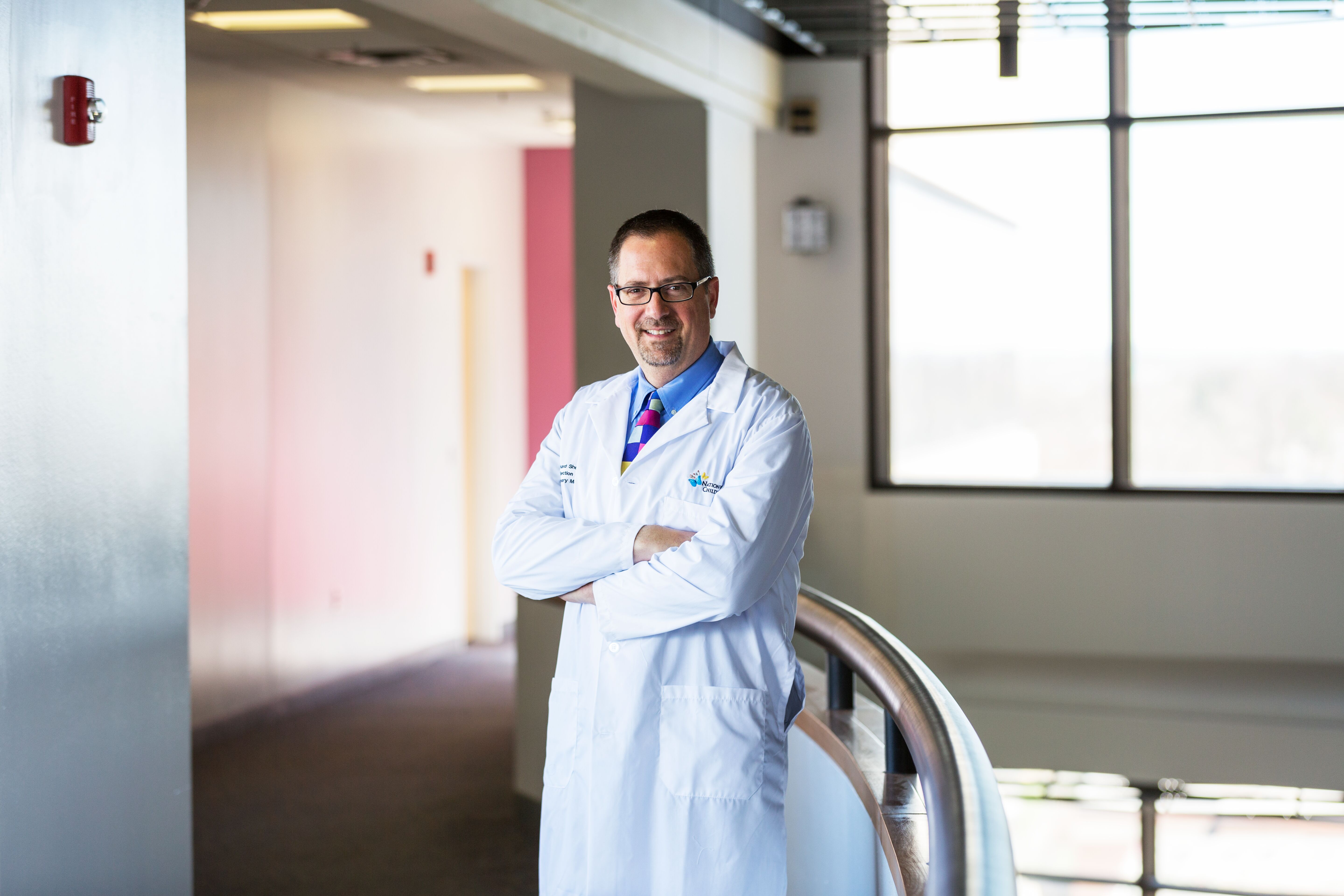 A physician smiling and standing confidently next to a railing in a hospital corridor.
