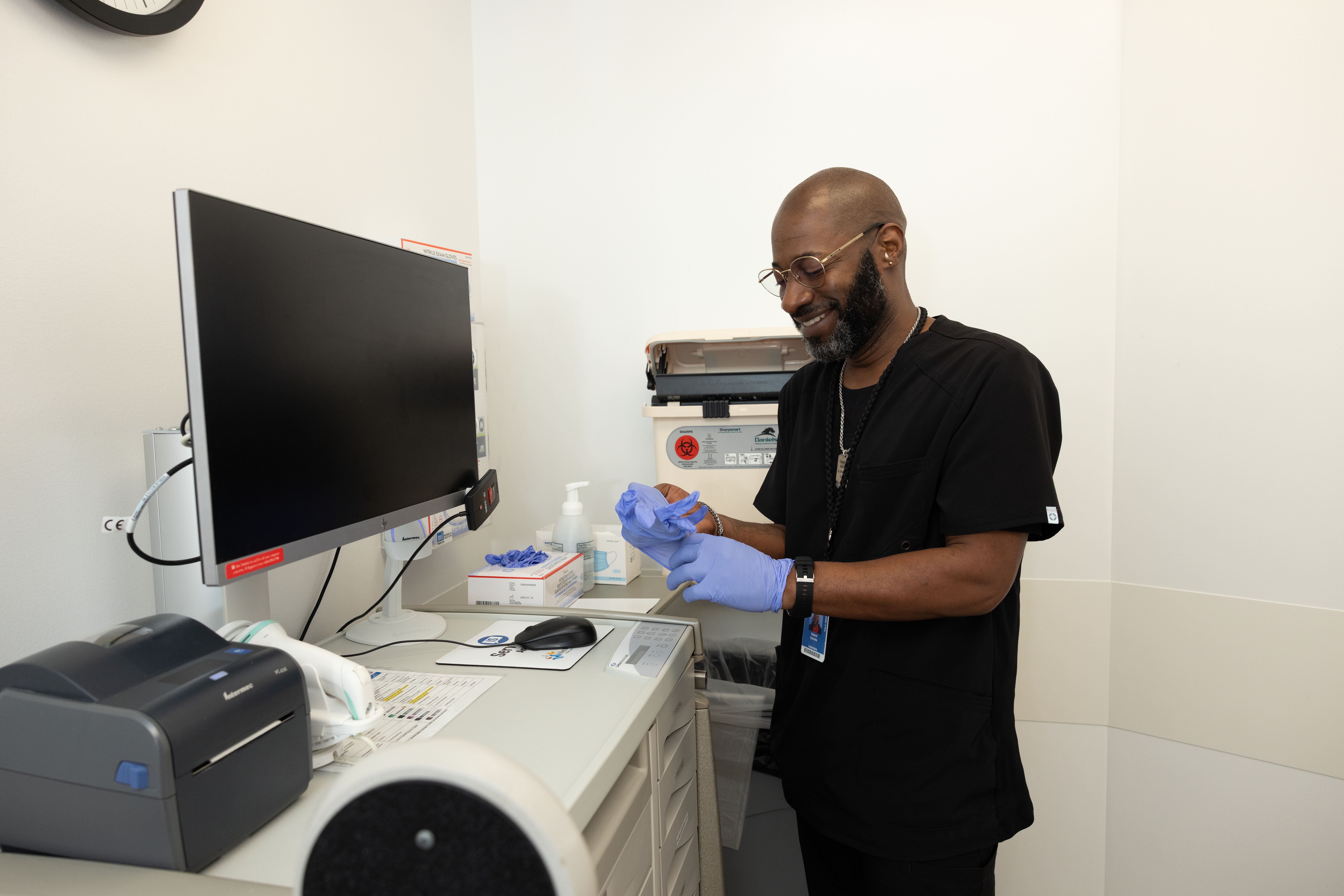 A healthcare worker in scrubs is preparing supplies while smiling in an examination room.