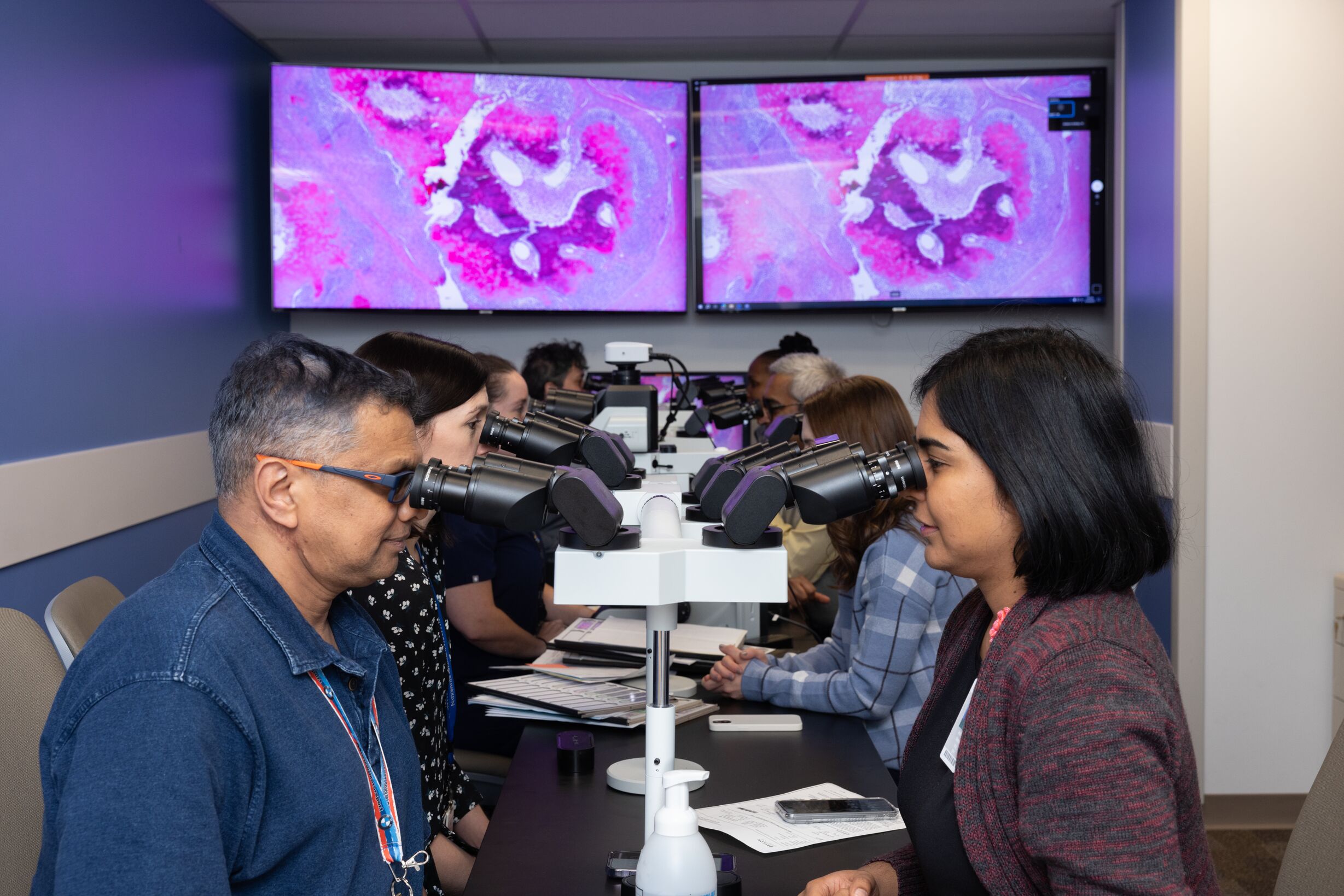 Two individuals use microscopes in a lab setting, with screens displaying magnified tissue samples in the background.