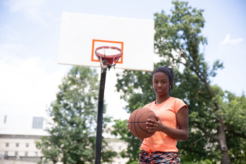 A teen girl holds a basketball with a serious expression. An outdoor basketball hoop is behind her on a sunny day.