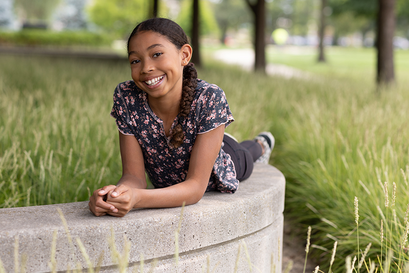 Smiling young girl lying on a low concrete wall surrounded by tall grass and trees.