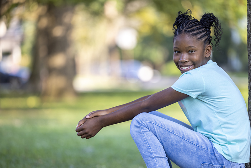 Smiling young girl sitting outdoors in a park with trees and grass in the background.
