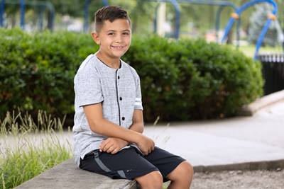 Boy sitting outside with bushes and a playset behind him. 