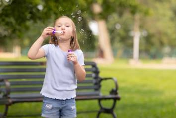 A young girl blowing bubbles in a park, with a bench visible in the background and greenery surrounding her.