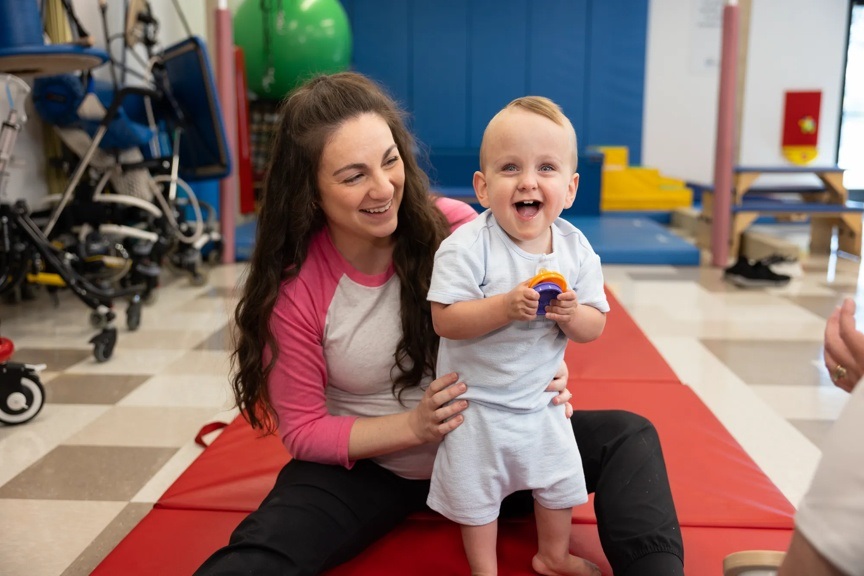 Physical therapist wearing a white and pink baseball shirt is holding a toddler while he is standing up. He is smiling at the camera and wearing white shorts and a white shirt.
