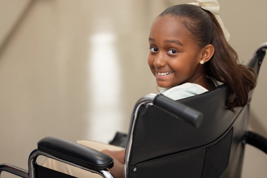 A young girl sits in a wheelchair and looks back over her shoulder, smiling warmly. She wears a light-colored outfit and a hair bow, appearing confident and cheerful in a bright indoor setting.