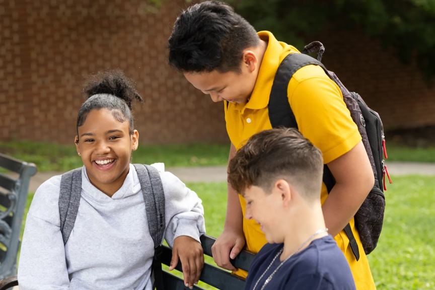 Three teenagers, two boys and a girl, are smiling and interacting in a park setting, likely sharing a fun moment together.